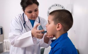 female doctor administers asthma inhaler to young boy in blue shirt