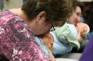 woman in pink shirt apply lips to ear of baby mannequin in classroom with others doing the same