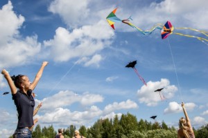 young adult female and middle aged adults flying brightly colored kites shaped like bats and triangles with long tails agains bright blue sky and fluffy wihite clouds