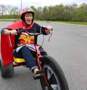 older adult woman riding oversized big wheel tricycle wearing a Wonder Woman costume